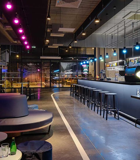 RGBW LED pendant lights in blue and magenta illuminate a bar and lounge in a bowling alley with cylinder downlighting for ambient mood lighting.