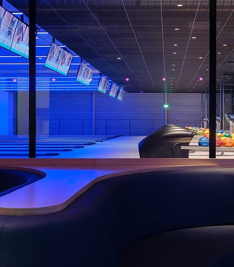 White linear recessed lighting provide general illumination over ball returns in a bowling alley with blue RGB lights illuminate the lanes.